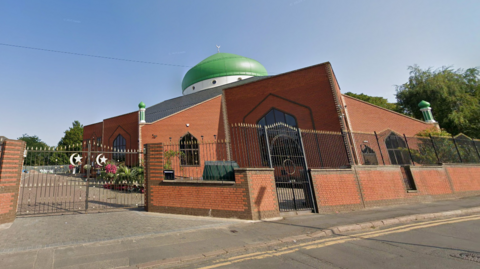 A Google Streetview image showing the exterior of the Central Mosque. It is a red brick building with a green copper dome on top.