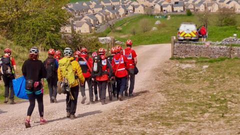 12 people in red hard hats and jackets walking along a gravel path towards a car. There are houses in the background. 