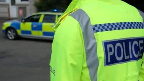 Police officer in hi-vis jacket taken from the back with a police car in the background.
