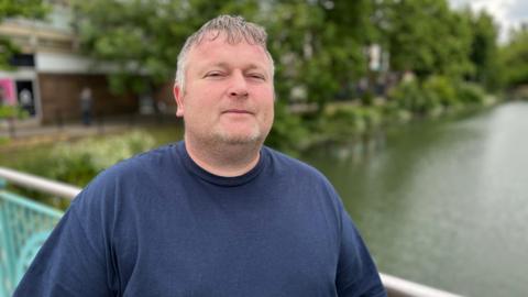 Liam Lathbury wearing a blue top and looking straight into the camera with a river in the background