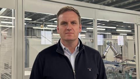 Cormac Diamond of Bloc Blinds has short brown hair and is staring into the camera. He is wearing a patterned shirt underneath a dark jacket. In the background are machines used to make blinds.