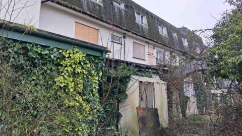 The outside of a derelict hotel in Guernsey which is boarded up and completely overgrown by greenery. 