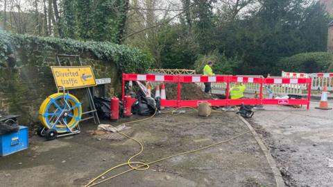 A road sign with a SGN fences blocking the road. There are two men wearing high-visibility jackets digging a hole. 