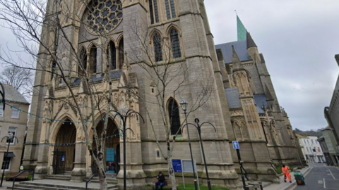 A google street view image of Truro Cathedral. It is the front of the Cathedral. The Cathedral is tall and made of a grey brownish brick. There are trees in front of it with string lights. 