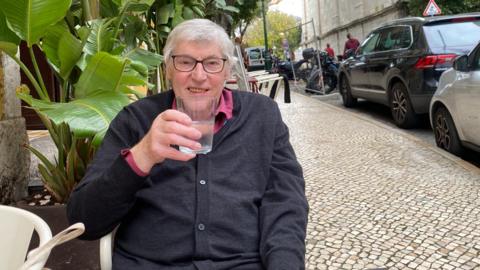 Jerry Cowhig sitting at a white table outside a restaurant. He is wearing a burgundy shirt underneath and buttoned black cardigan. He has white hair and black framed glasses. He is holding up a glass of water and smiling at the camera. The pavement beside him is covered in pale brown stones and there are cars parked on the road. Behind Jerry is a tropical plant in a large pot.