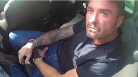 Police bodycam footage showing Paul Farrelly, 46, who has short brown hair and is wearing a dark blue t-shirt and jeans sitting in the back of a vehicle. He is in handcuffs and is looking up at the officer.