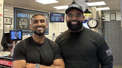 Two men smiling into the camera in a white-tiled butcher's shop. One has short hair and a slight beard. The other has a cap on with glasses and a bushy beard. 