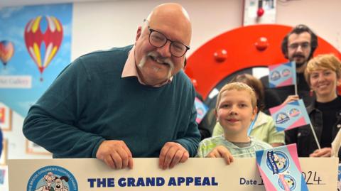 Michael Carter (left), holds a giant cheque with Harry. There are people behind them smiling and holding blue and pink Grand Appeal flags. 