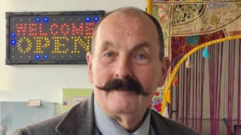 A balding man with a thick brown handlebar mustache is wearing a tweed jacket and standing in from of a coloured sign which reads Welcome Open