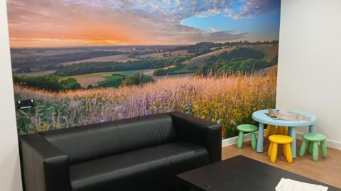 A room inside the SARC which has a black sofa, a black coffee table and a children's set of a table and four chairs standing on a wood-style vinyl floor. There is a large photograph filling the wall behind the furniture which shows a rural landscape, a field of wild flowers and rolling hills under a sunset with dramatic clouds.   
