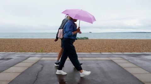 Two people walk along a seafront, one of them holding an umbrella, with a pebble beach and the sea in the background and a grey sky overhead
