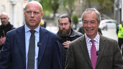 Dan Thomas is wearing a blue suit and pokadot tie with a blue shirt next to Nigel Farage, who is wearing a chequered suit, lightly chequered shirt and purple tie.