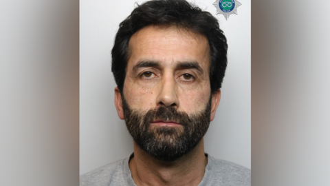 A police mugshot showing a man with a dark beard and short black hair.