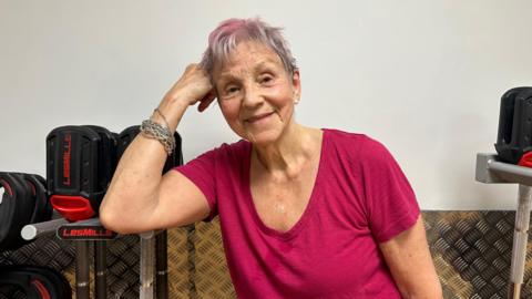 Lynne Thompson poses with her head resting on her arm. She is wearing a dark pink top and has grey and pink short hair.