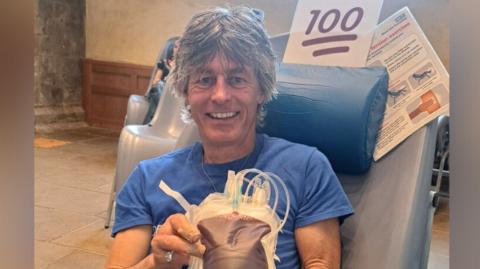 A man in a blue T-shirt smiles as he holds up a bag of blood. He is sat on a blood donation chair.