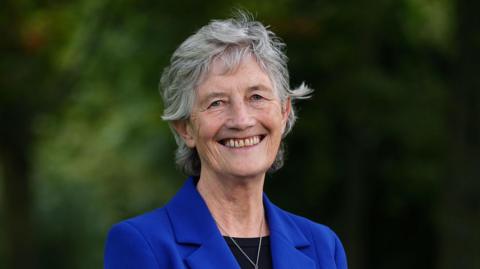 A close-up image of Catherine Connolly smiling at the camea. She is an older woman, who has short grey hair. She is standing in a park, but the trees behind her are blurred. She is wearing a cobalt blue blazer jacket, with a black shirt and a silver necklace.