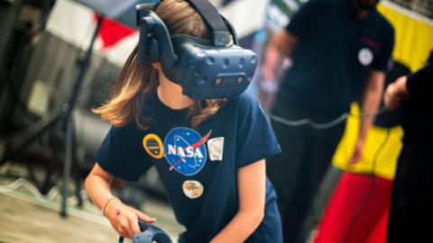 The picture shows a young person taking part in a virtual reality activity at what appears to be a science, technology or space‑themed event. In the centre of the image, the person is wearing a virtual reality headset that fully covers the eyes and upper face, with a thick strap running over the head. A handheld VR controller is visible in one hand. They are wearing a dark T‑shirt with a large NASA logo on the front, along with several circular patches or badges.