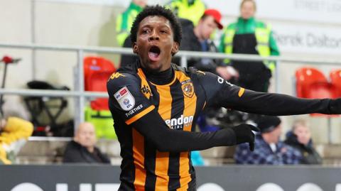 Jaden Philogene, wearing Hull's black and gold striped top, celebrates a goal as he shouts with an open mouth and swings both arms out to the left of him