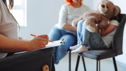 A child in a therapist's office sitting on a chair with a teddy bear sat next to a woman in blue jeans. the therapist is sitting opposite them holding a file and a pencil 