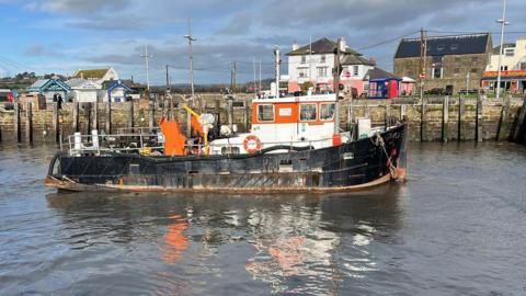 Side view of dredging vessel Doonhammer in Bridport harbour. The boat is about 15 metres long and has a black hull with an orange and white cabin. In the background is the stone harbour wall lined with wooden mooring posts. Along the quayside are lines of wooden takeaway huts and some Victorian-era buildings, including a brick warehouse, a white and pink painted pub and some terraced houses and shops.