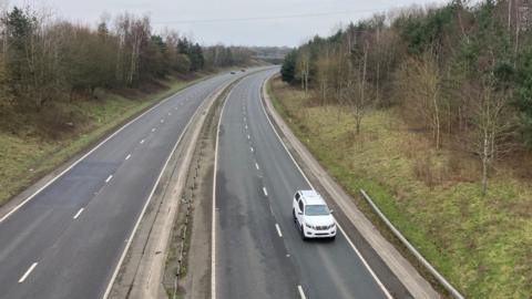 A dual carriageway, with trees and a grass verge on either side of the road. There is a white car travelling towards the bottom of the image. 