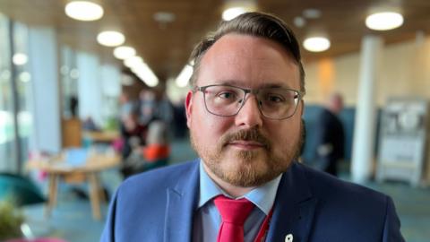 A man with glasses and a beard wearing a blue suit and shirt with a red tie looking at the camera with a neutral facial expression. A reception area in an office building is out of focus behind him.