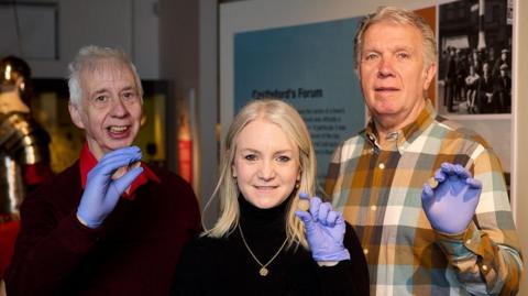 Two men with grey hair and one woman with blonde hair hold coin shaped white discs. They are all wearing blue gloves.