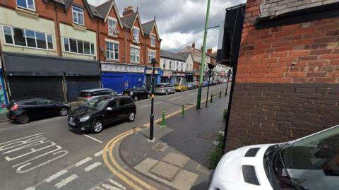 A street with commercial premises running along the left side. There is a row of cars parked on the street. To the right is a red brick building