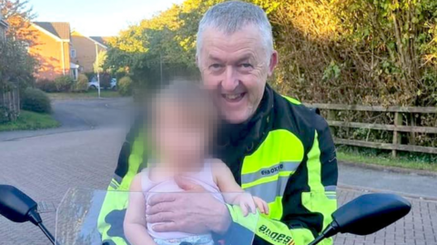 Motorcyclist, Nigel Osler, sat on a stationary motorbike in a driveway, carrying a baby whose face is blurred
