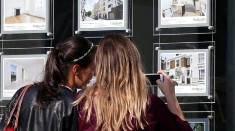 Two young women with their backs to the camera take a photo of an advert for a house in an estate agent's window. The woman on the left has black hair tied in a ponytail and a black leather jacket. The woman on the right has shoulder-length blonde hair and is holding her phone up to take a photo. 