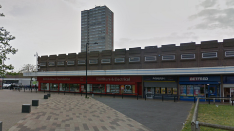 A google street view image of Salford Shopping Precinct. There is a British Heart Foundation furniture shop, an Admiral, Betfred, and a tower in the background with the words 'Salford Shopping City' written on the side.