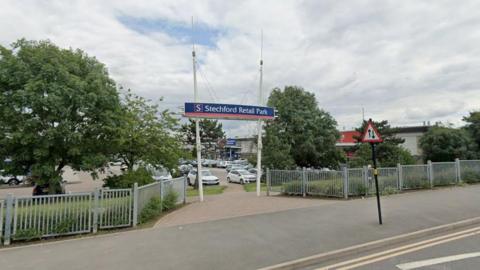 A Google street view image showing the entrance to a retail park on a pavement next to a main road. A blue, red and white sign above the entrance says "Stechford Retail Park". There are grey fences and trees either side. Beyond the entrance there is a car park and a line of large shops. 