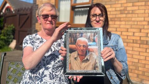 Two women hold up a silver framed picture of an older man.
The woman on the left, Susan Holmes, is wearing a black and white floral patterned top and dark glasses and has short blonde hair while the woman on the left, Lisa Jones, has long dark hair and is wearing a denim jacket and glasses. The man has short white hair and is looking directly at the camera while wearing a striped shirt. Behind them you can see the wall and window of a house.