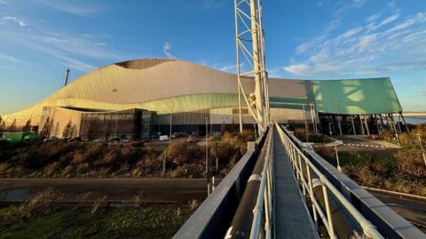 A large, modern building with a distinctive wave-like roof design, combining metallic and green-tinted panels. The structure appears industrial or possibly a sports or event venue, with a sleek, contemporary style. In the foreground, a metal walkway with railings extends toward the building, creating strong leading lines. To the right of the walkway, a tall, white structural tower rises vertically, adding to the industrial aesthetic. The surrounding area includes a road, landscaped greenery, and several parked vehicles near the entrance. The sky above is clear with scattered clouds, bathed in warm sunlight, giving the scene a bright and crisp appearance.