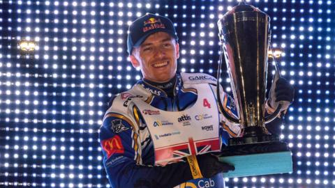A male wearing a speedway biker outfit holding a large silver trophy while smiling. He is standing in front of a wall covered in small lights.