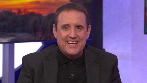 Peter Kay sits on a green sofa, he wears a black knitted polo shirt under a black jacket. He has short dark hair and smiles widely at the camera