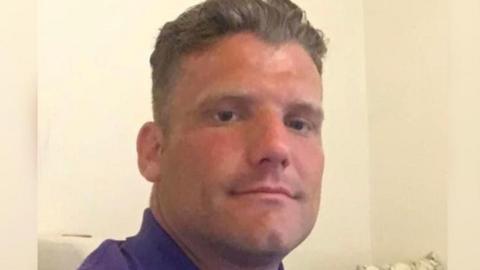 Sheldon Jeans has combed back dark hair and a purple polo shirt and is sat in front of a cream wall.