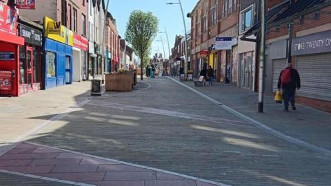 A high street which is lined with shops on either side. The shop in the foreground on the right is boarded up and there is a "to let" sign above the shop next to it. The pedestrianised street has planters and trees down the middle. There is a man walking away from the camera on the right and people visible in the distance.