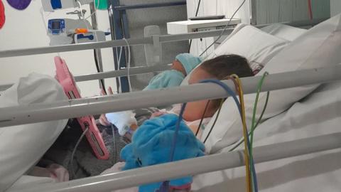 Myah lying in a hospital bed watching a tablet. She has various tubes and wires attached to her 