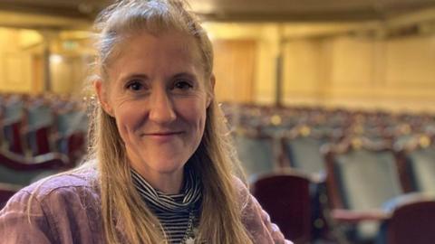 Head-and-shoulders shot of Kerry Vasiliou, sitting in a theatre flooded with  light. She is smiling and has long blonde hair tied back off her face.