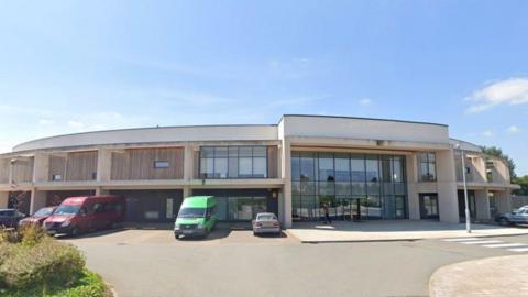 The main entrance to the school includes a two storey glass panelled section. Several cars and vans are parked in front of the building.