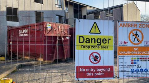 A metal fence in front of a grey building with no windows in. There is a red tip in front of the building. On the metal fence are danger signs that say "no unauthorised access" and "no entry beyond this point." The sky is blue. 