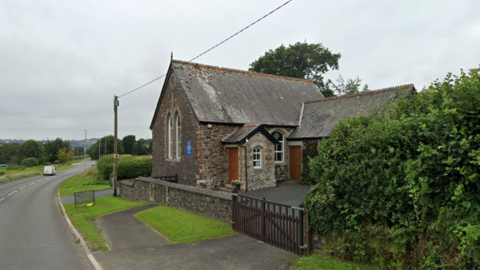 Rural scene along the A388 road. Vehicles, including a car and a van, are traveling on the left side. On the right side, there's an old stone church with a slate roof, arched windows, and wooden doors, with a small gated entrance. The area is lush with greenery, including bushes and trees, with a grey, cloudy sky behind.