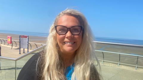 A closeup photo of a woman with blonde hair and black rimmed glasses, smiling to the camera in front of Blackpool Prominade