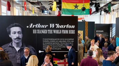 A large room with people looking at exhibits. A large board bears a black-and-white picture of Arthur Wharton with the words: "He Changed The World From Here" on it. There are flags and football shirts hanging from the ceiling.