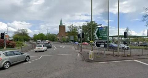 A modern church is in the background of a road juction with signs to London and Watford, and cars travelling around a bend.