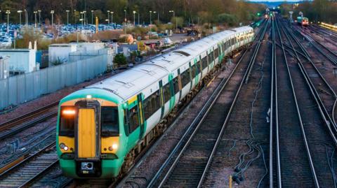 A green train on a railway line. A large car park is to the right of the tracks.