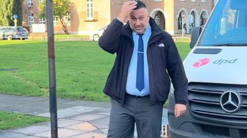 The image shows a man in a suit, black jacket, blue shirt and dark blue tie. He is trying to cover his face with his hand. There is a white van to his left and a large building behind him.
