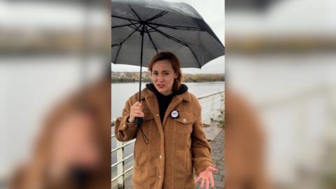 A girl with auburn hair is wearing a light brown coat, a black hooded jumper underneath and silver hoop earrings. She is holding a black umbrella and is standing in front of a grey river underneath grey skies in Glasgow.