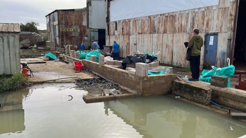 Men working on the Arun Shipyard to improve its flood defences, with a brick wall and water in the foreground 